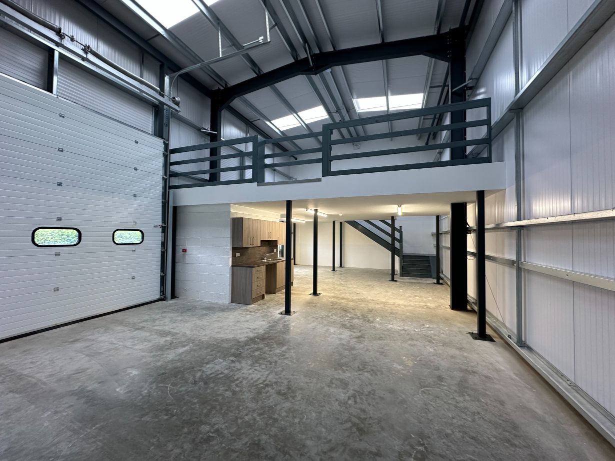 Interior of an empty industrial unit with a large roller door, mezzanine level, and kitchenette area.