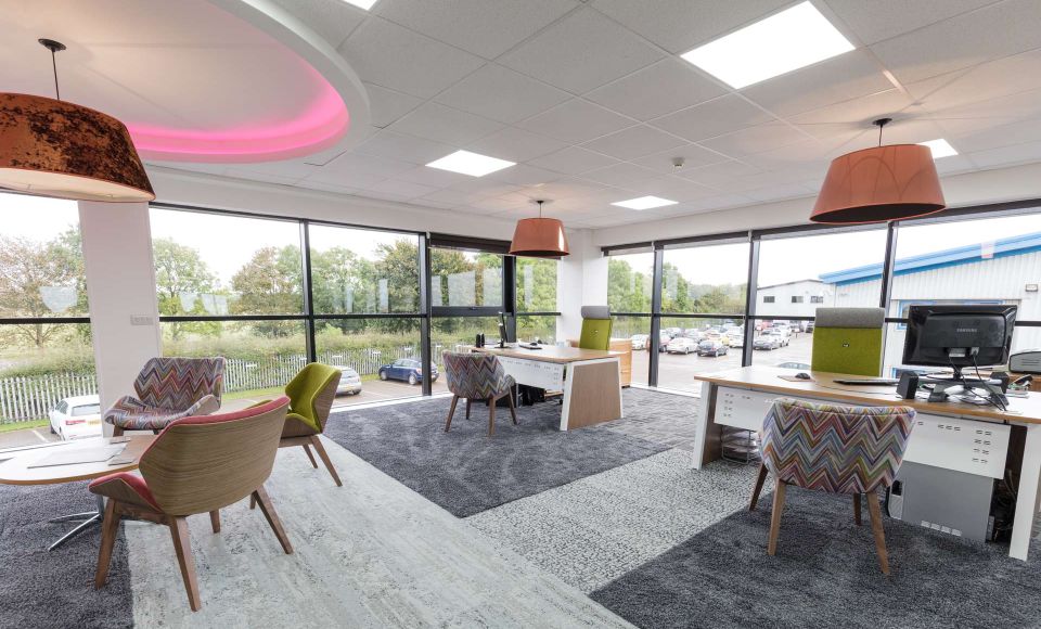 A modern office, decorated with dark grey carpets, chairs, desks and computers. The large glass windows overlook an occupied carpark. 